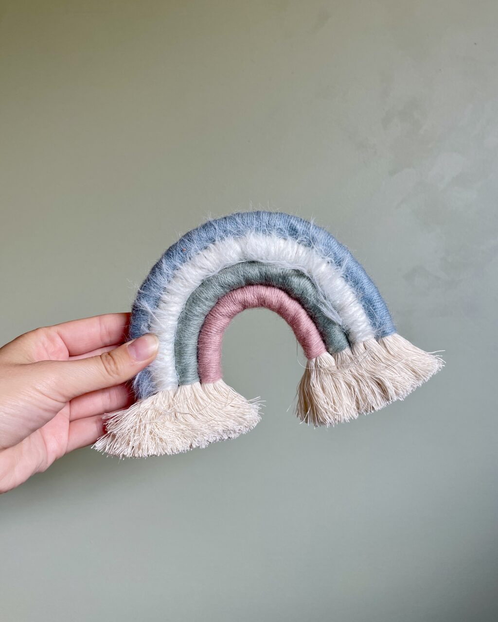 Håndlavet regnbue i pasteltoner hængende på en lys væg – lavet af bomuldsreb og garnrester i dusty rosa, salviegrøn, off-white og støvet blå. En unik vægdekoration med et blødt og legende udtryk.