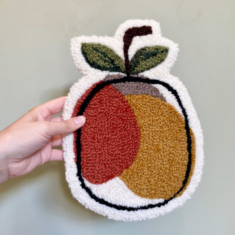 Tuftet vægophæng formet som et æble i efterårsfarver – varme nuancer af orange, karrygul og brun. Håndlavet, unikt dekorativt kunstværk til væggen, ca. 20 x 29 cm.